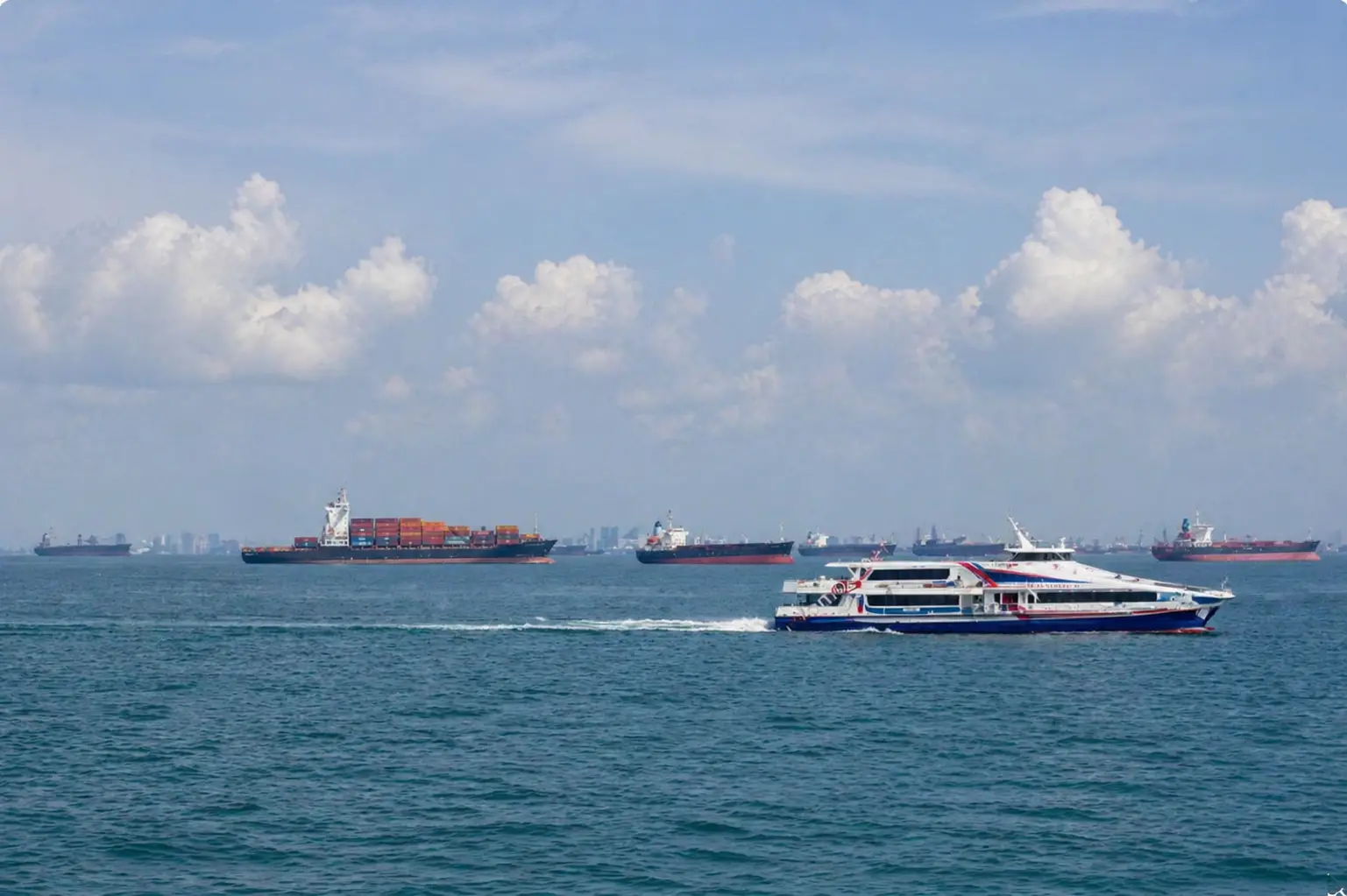 Passenger ferry crossing the Strait of Malacca with multiple cargo ships on the horizon under hazy skies