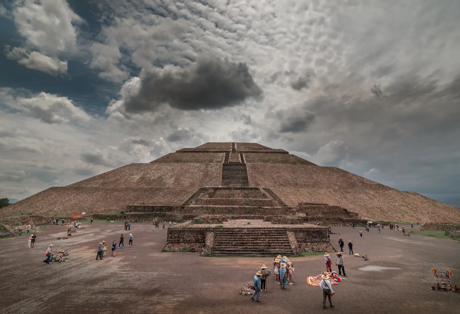 Tourists and street vendors gather at the base of the Pyramid of the Sun at Teotihuacan, Mexico, under a dramatic cloudy sky