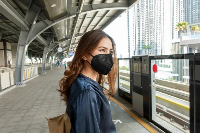 Female traveller wearing black anti-pollution face mask waiting on urban train platform in modern city with high-rise buildings