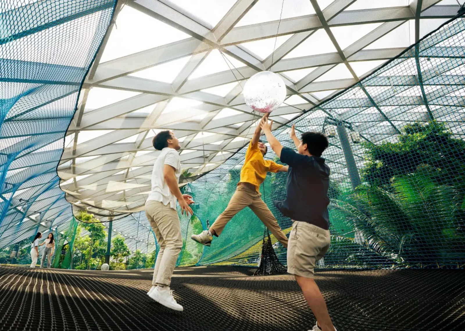 Jewel Changi Airport: Key Highlights Of This Iconic Lifestyle Destination (Part 2) 5 Three visitors jumping and playing with a ball on the Bouncing Net attraction at Canopy Park, Jewel Changi Airport, with tropical greenery and the glass dome structure visible behind them.