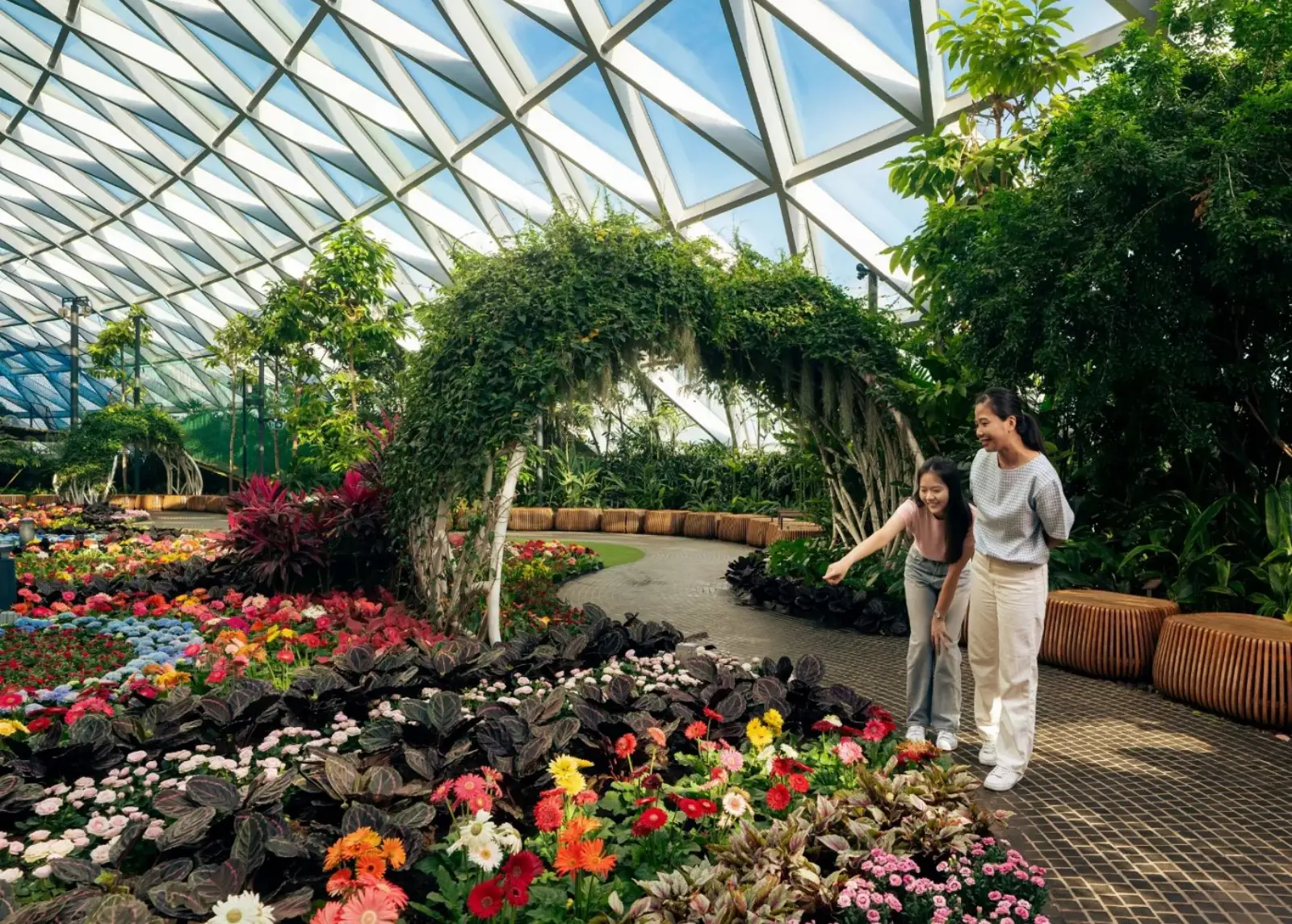 Jewel Changi Airport: Key Highlights Of This Iconic Lifestyle Destination (Part 2) 6 A woman and child walking along a path through the colourful seasonal floral displays of the Petal Garden at Canopy Park, Jewel Changi Airport, with a green leafy arch and glass dome overhead.