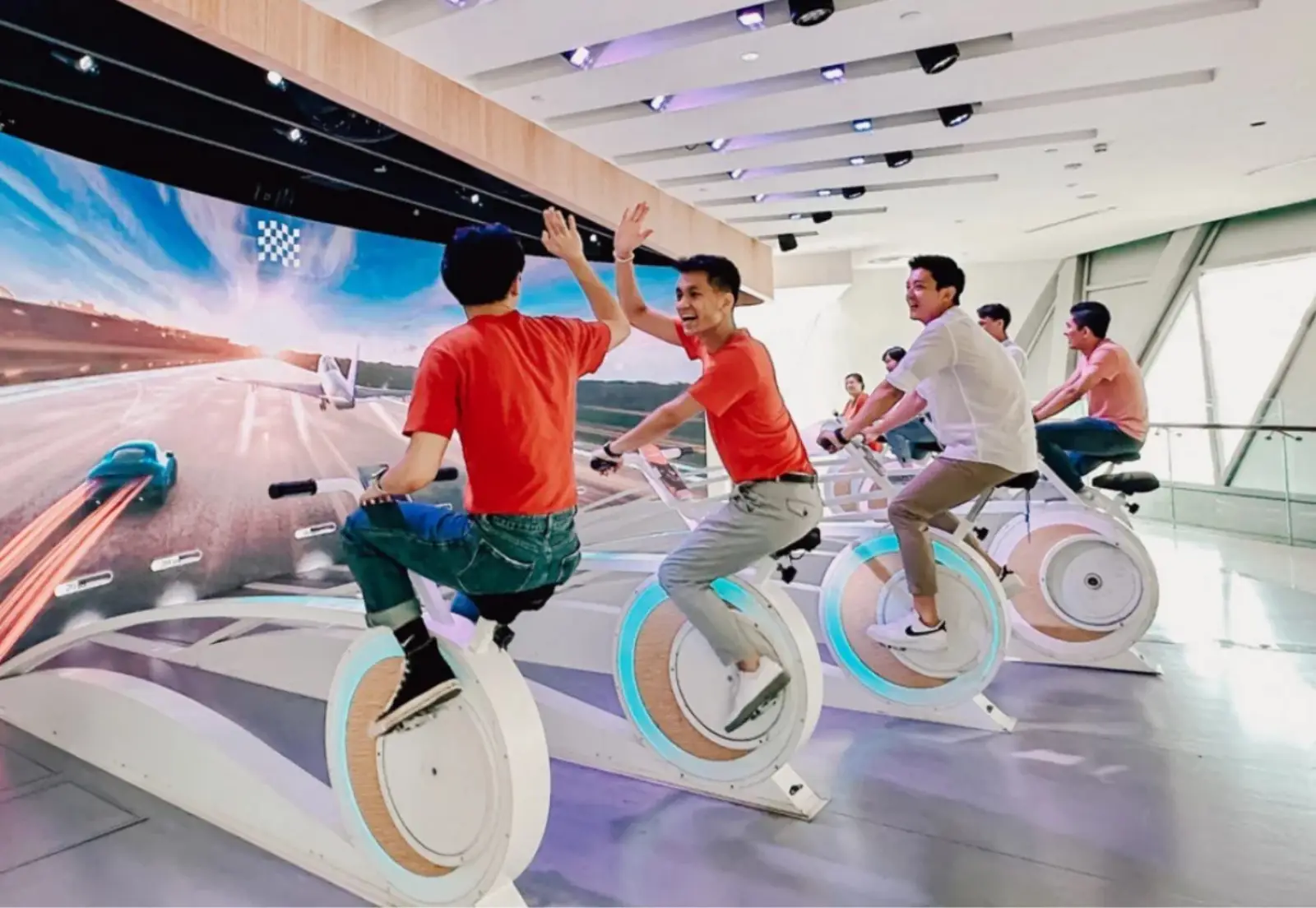 Jewel Changi Airport: Key Highlights Of This Iconic Lifestyle Destination (Part 2) 8 Visitors riding stationary bikes and high-fiving during the Amazing Runway multiplayer game at the Changi Experience Studio, with a large screen showing an airport runway race behind them.