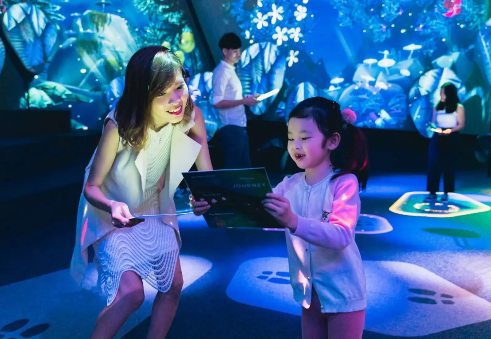 Jewel Changi Airport: Key Highlights Of This Iconic Lifestyle Destination (Part 2) 9 A woman and young child looking at a journey card together inside the Garden of Harmony at the Changi Experience Studio, surrounded by immersive blue-lit projections of tropical plants and flowers.