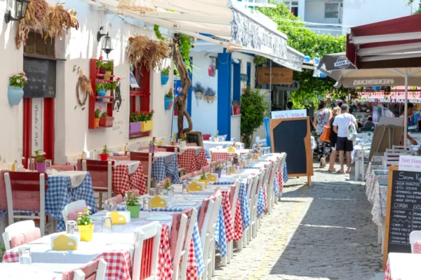 Outdoor restaurant tables with red and blue checked tablecloths lining a narrow cobbled street in Bozcaada, Turkey, with tourists walking past whitewashed buildings decorated with colourful flower pots