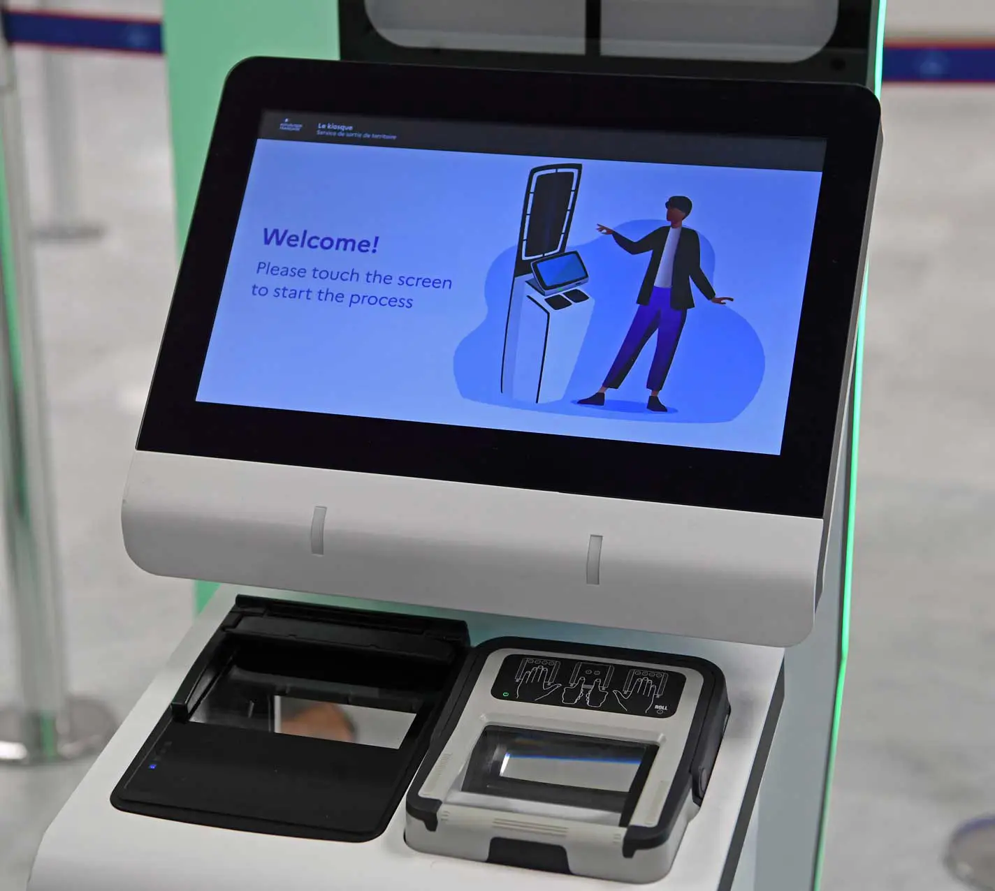 A white self-service EES registration kiosk at an airport border control point, featuring a touchscreen displaying a welcome message and an animated figure, alongside a fingerprint scanner and document reader