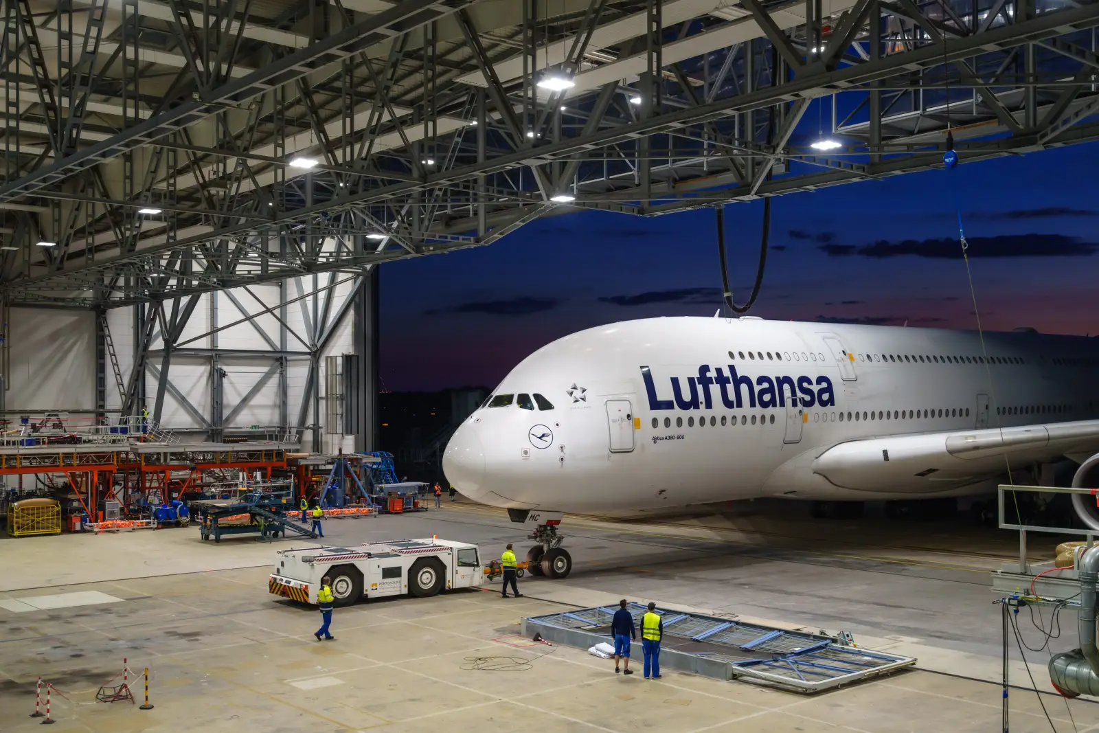 A Lufthansa Airbus A380-800 wide-body aircraft inside a large maintenance hangar at dusk, with ground crew in high-visibility jackets working around the aircraft and a tow vehicle positioned near the nose gear