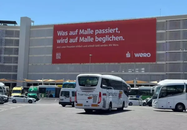 A large red advertising banner with white text covers the facade of the car park building at Palma Airport, Mallorca. The German-language slogan reads "Was auf Malle passiert, wird auf Malle beglichen" with the Sparkasse and Wero logos. Coaches and vehicles are visible in the foreground.