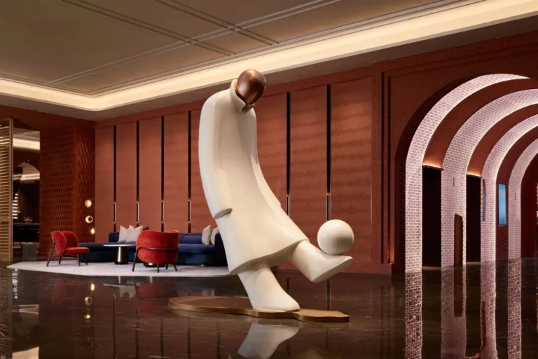 The grand lobby of Radisson Blu Hotel, Shanghai Stadium, featuring a large white sculptural figure with a ball at its feet referencing sport, set against terracotta-toned wall panels, arched passageways, and a seating area with navy and red upholstered furniture on a polished marble floor.