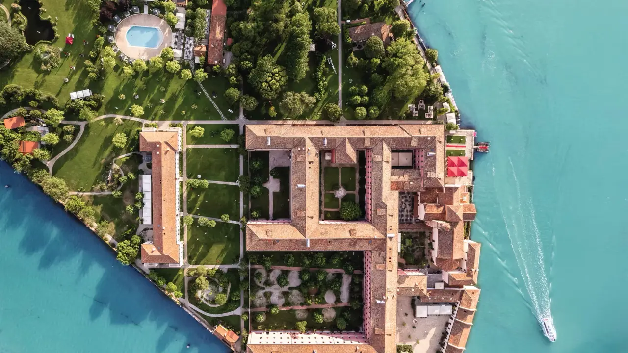 The Most Anticipated Hotel Openings In Venice In 2026 5 An aerial view of San Clemente Palace Venice showing the hotel's private island set within the calm waters of the Venetian lagoon. The historic complex features terracotta-roofed monastery buildings, a domed Romanesque church, and extensive Mediterranean gardens with tall cypress trees, manicured lawns and courtyards. An outdoor swimming pool is visible among the greenery, and the Venice city skyline with its distinctive domes and bell towers is visible in the distance across the lagoon under a clear blue sky.