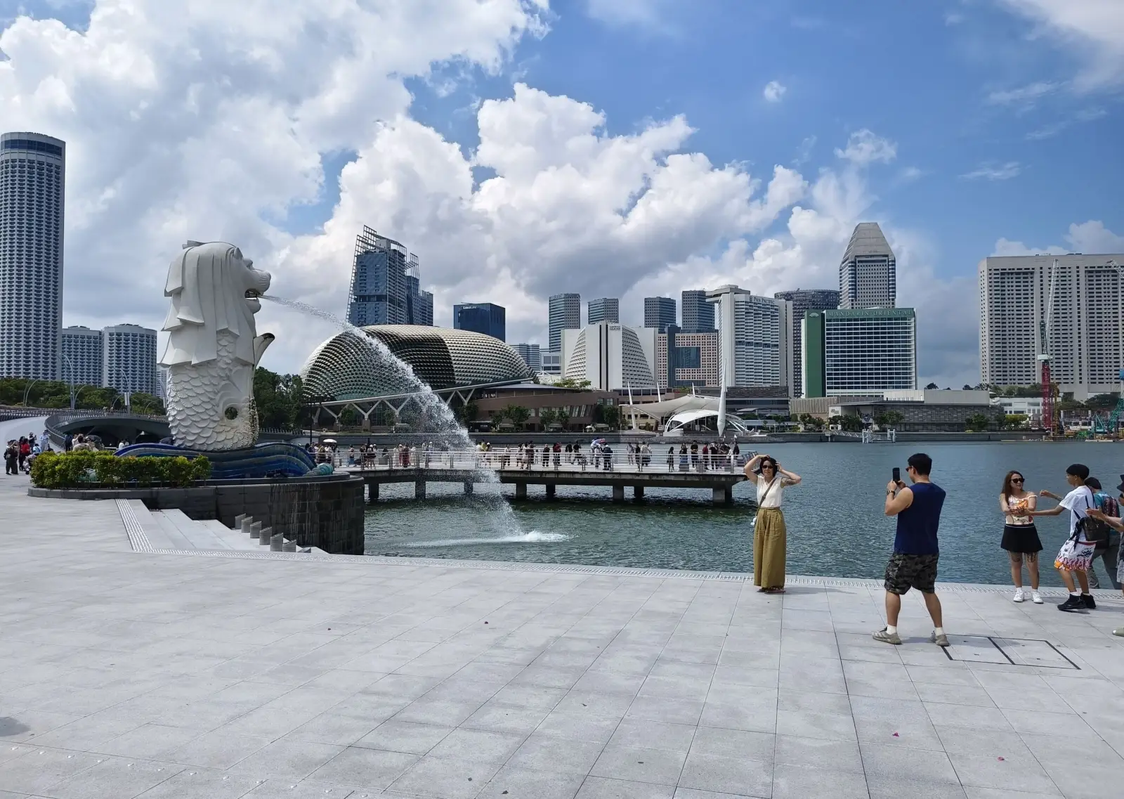 Tourists posing for photographs near the Merlion fountain statue at Marina Bay, Singapore, with the Esplanade Theatres on the Bay and the city skyline visible across the water under a partly cloudy sky