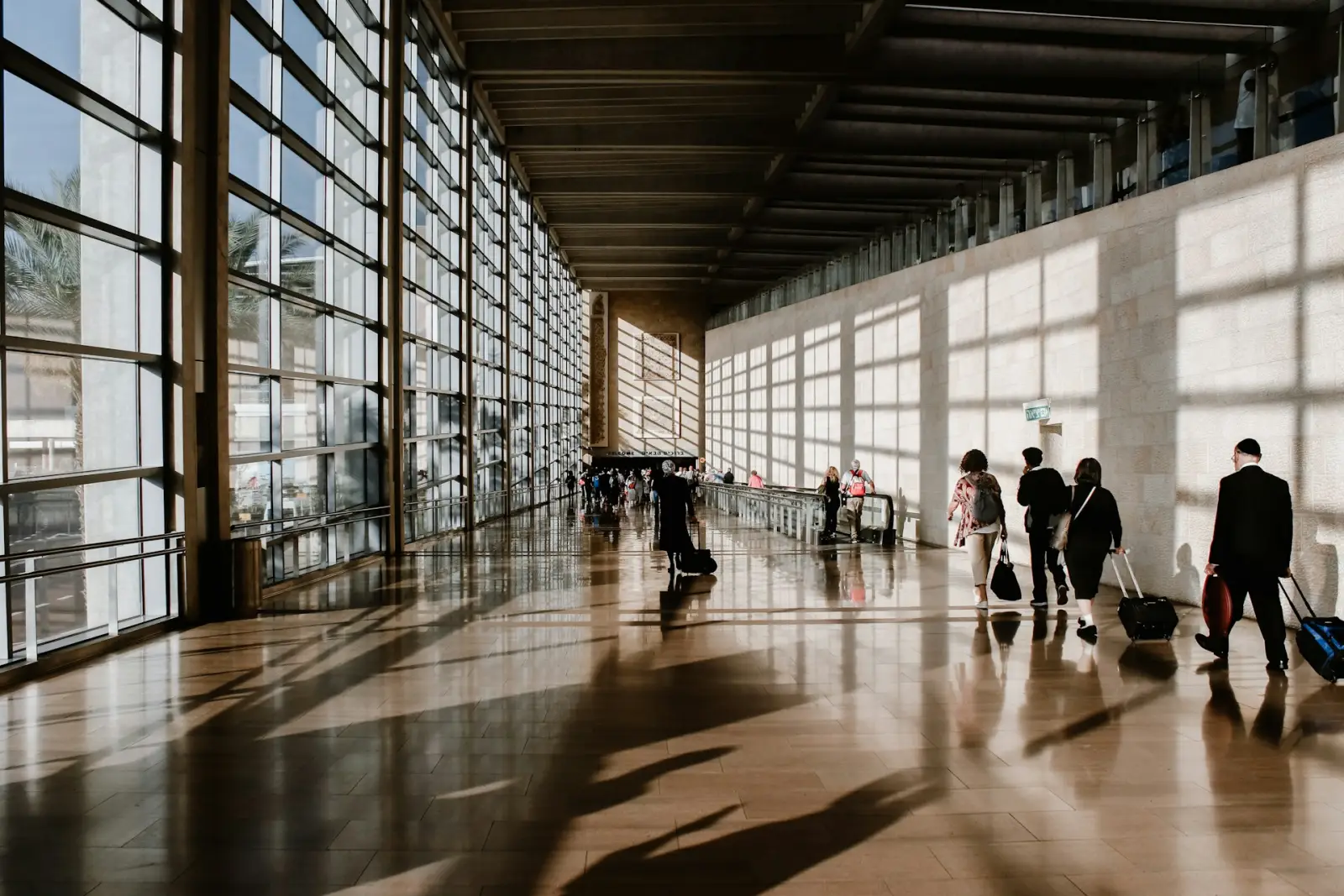 Travellers walk through a spacious airport lounge corridor with floor-to-ceiling windows and warm afternoon light.