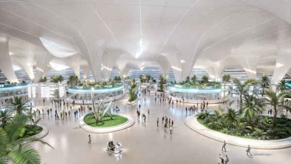 An architectural render of the entrance hall at Al Maktoum International Airport showing a vast, light-filled terminal with sweeping white arched ceilings, indoor palm trees and tropical greenery arranged in circular garden islands, passengers walking across a wide open concourse, and an autonomous passenger vehicle in the foreground