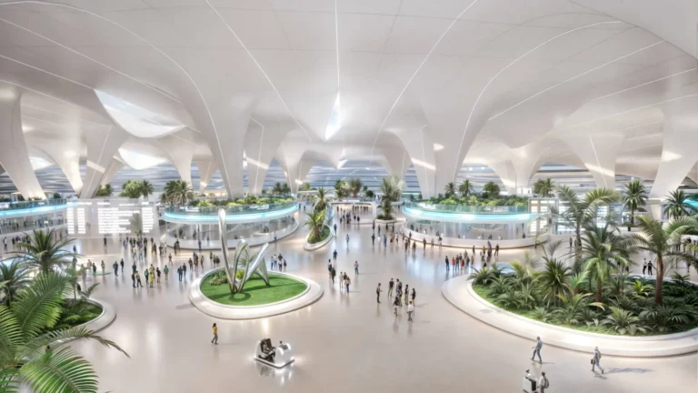 Aviation 17 An architectural render of the entrance hall at Al Maktoum International Airport showing a vast, light-filled terminal with sweeping white arched ceilings, indoor palm trees and tropical greenery arranged in circular garden islands, passengers walking across a wide open concourse, and an autonomous passenger vehicle in the foreground