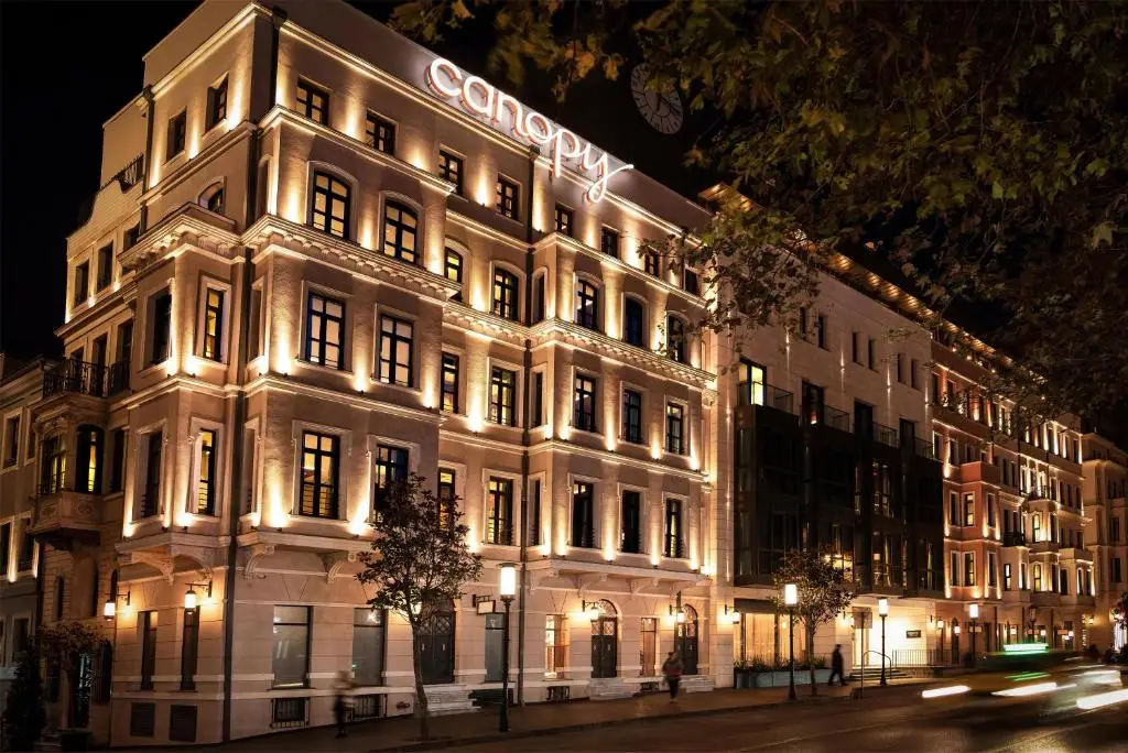 Istanbul Leads Europe In New Hotel Openings For 2026 14 The illuminated neoclassical facade of Canopy by Hilton Istanbul Taksim at night, featuring warm golden uplighting across five floors of arched windows and ornate stonework, with the Canopy brand name in neon lettering on the roofline, street trees in the foreground and passing traffic leaving light trails on the cobblestone street below