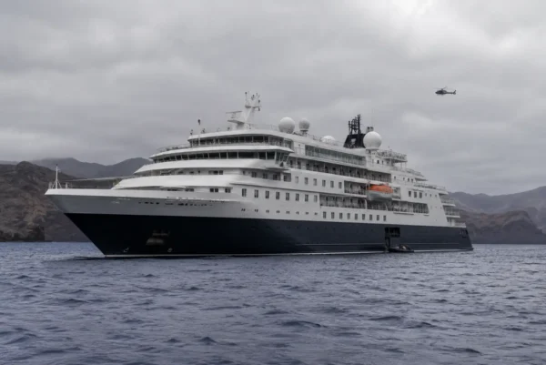 Modern expedition cruise ship anchored off the Canary Islands under grey skies with a rescue boat and distant helicopter nearby.