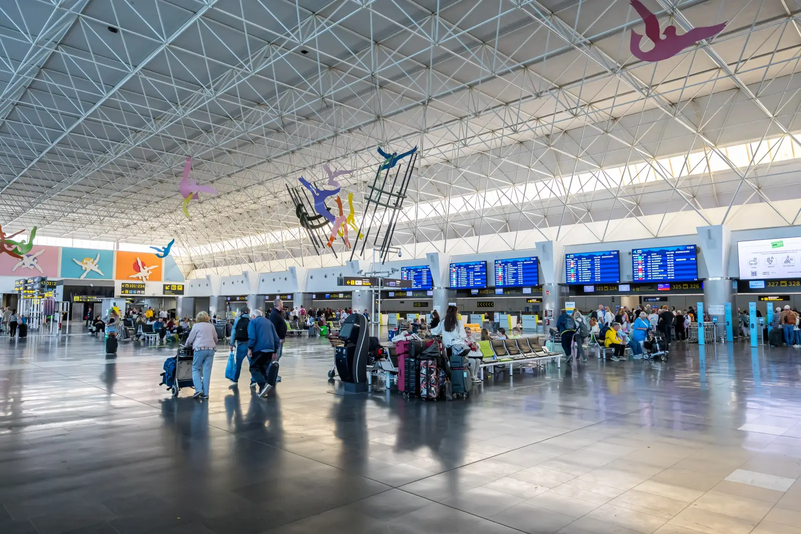 Passengers wait near check-in counters inside Gran Canaria Airport terminal in Spain with flight information screens overhead.