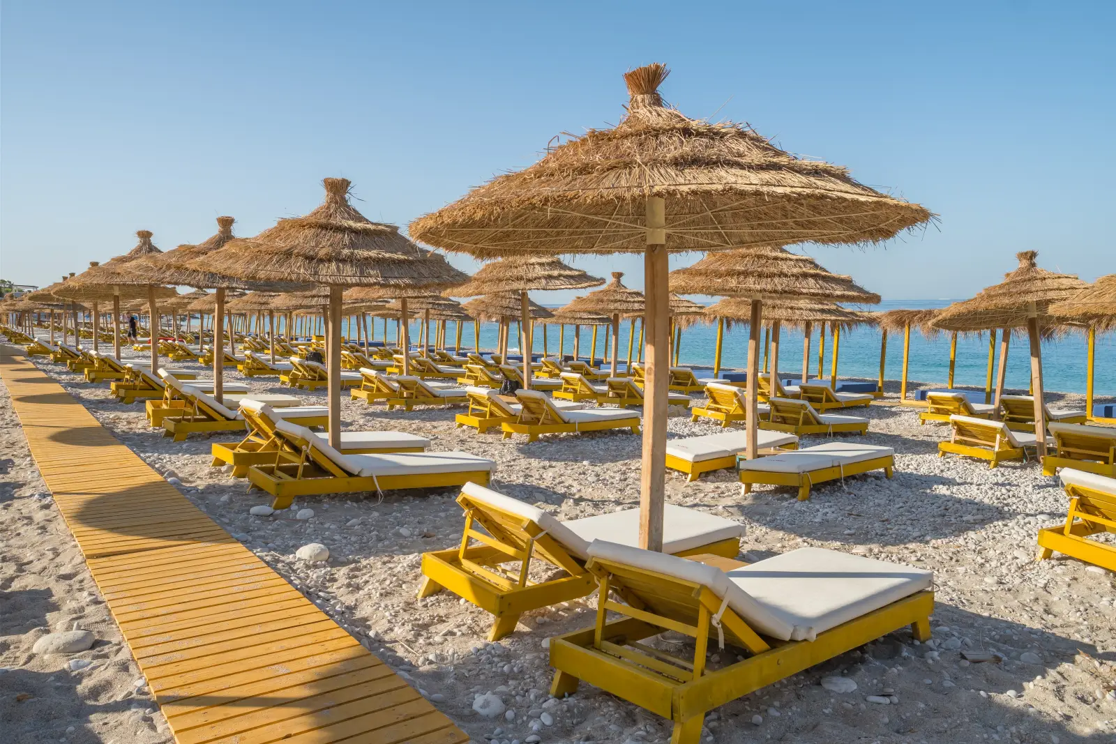 Rows of reserved sunbeds and straw umbrellas lined up on a beach resort in Greece under clear blue skies.