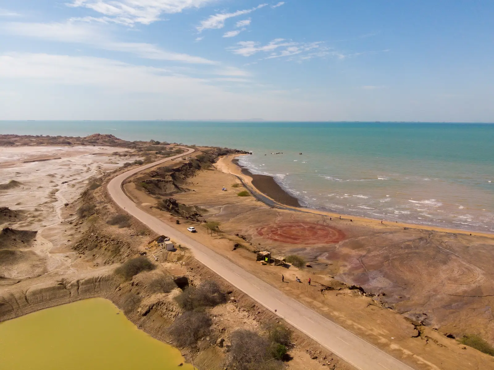 Aerial view of Hormuz Island in Iran showing the island’s red mineral-rich beach, coastal road and turquoise waters along the Strait of Hormuz under a clear blue sky.