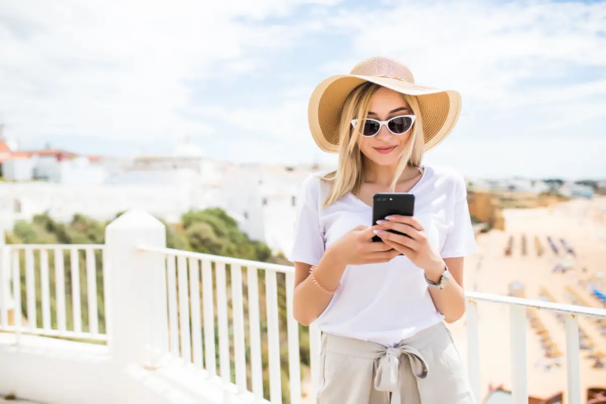 Woman using a smartphone with an eSIM while traveling in Italy, standing on a sunny terrace overlooking a coastal town.