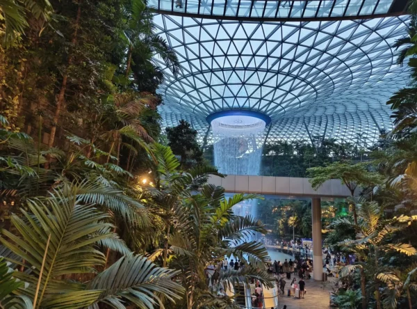 The Rain Vortex indoor waterfall at Jewel Changi Airport, surrounded by lush tropical trees and palms under a geometric glass dome, with visitors gathered below.