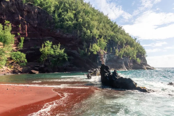 Kaihalulu Red Sand Beach on Maui features striking red volcanic sand, turquoise water and rugged lava rock formations.