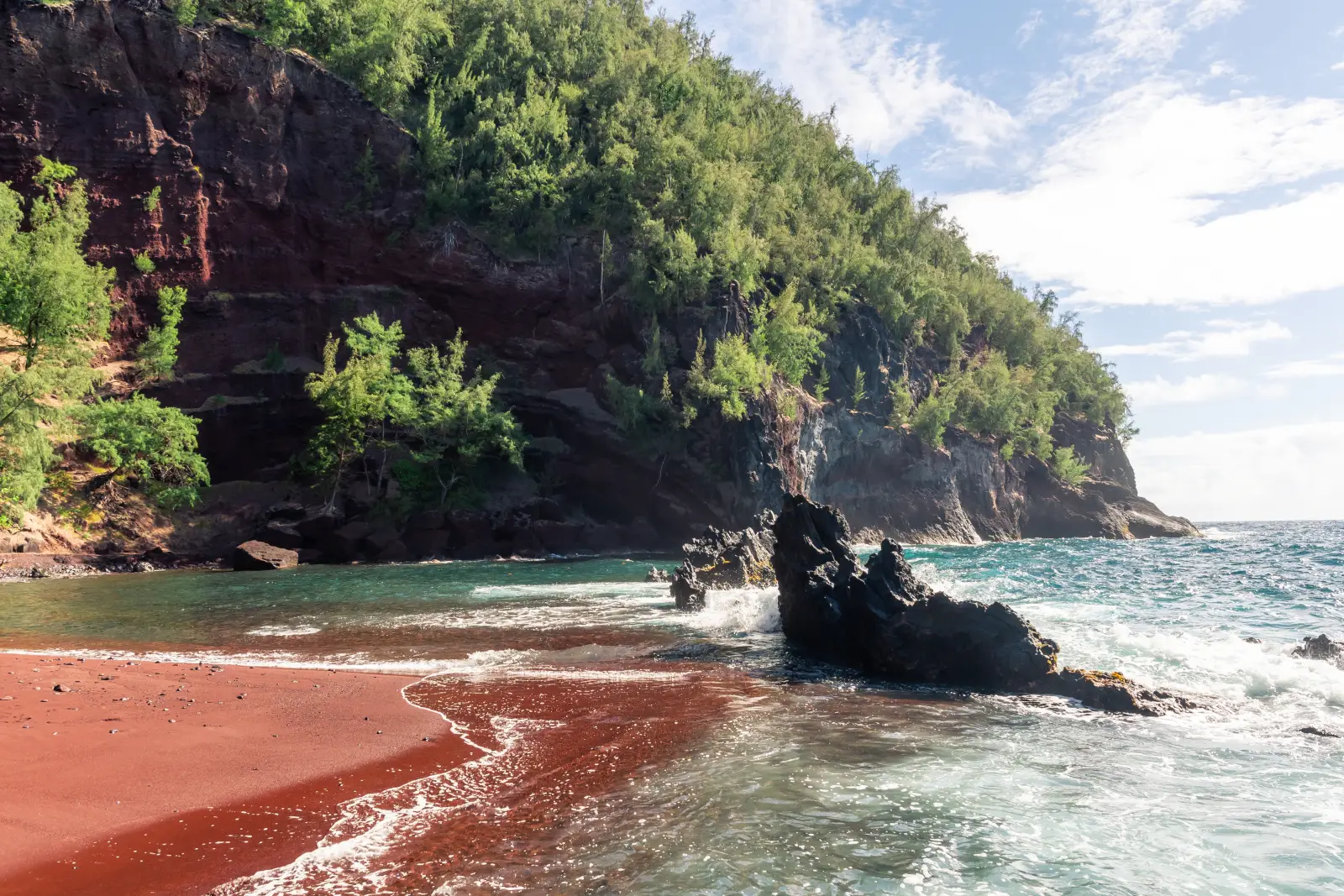 Kaihalulu Red Sand Beach on Maui features striking red volcanic sand, turquoise water and rugged lava rock formations.