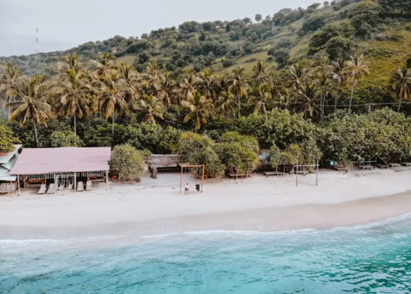 Aerial view of a secluded beach in Lombok, Indonesia, featuring calm turquoise water lapping onto white sand, dense tropical palm trees and lush green hills in the background, with small traditional beachside shelters and two visitors standing on the shoreline