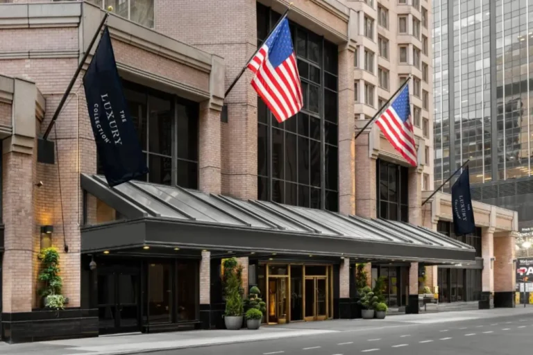 Street-level entrance of The London, a Luxury Collection Hotel, on West 54th Street in Midtown Manhattan, with American flags and branded banners above the canopied doorway