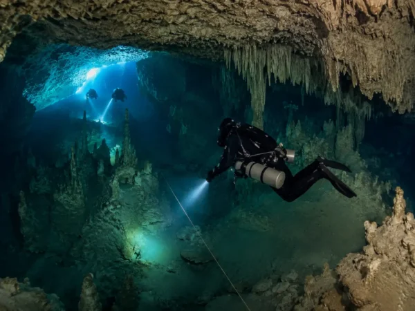 Technical scuba diver exploring a deep underwater cave system with flashlight beams illuminating rock formations during a cave diving expedition.