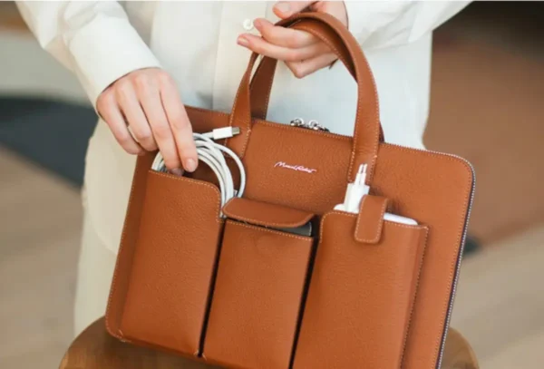 A person in a white blazer holding a tan full-grain leather structured laptop bag by Marcel Robert, with multiple front pockets organising a USB cable, charger and other travel accessories