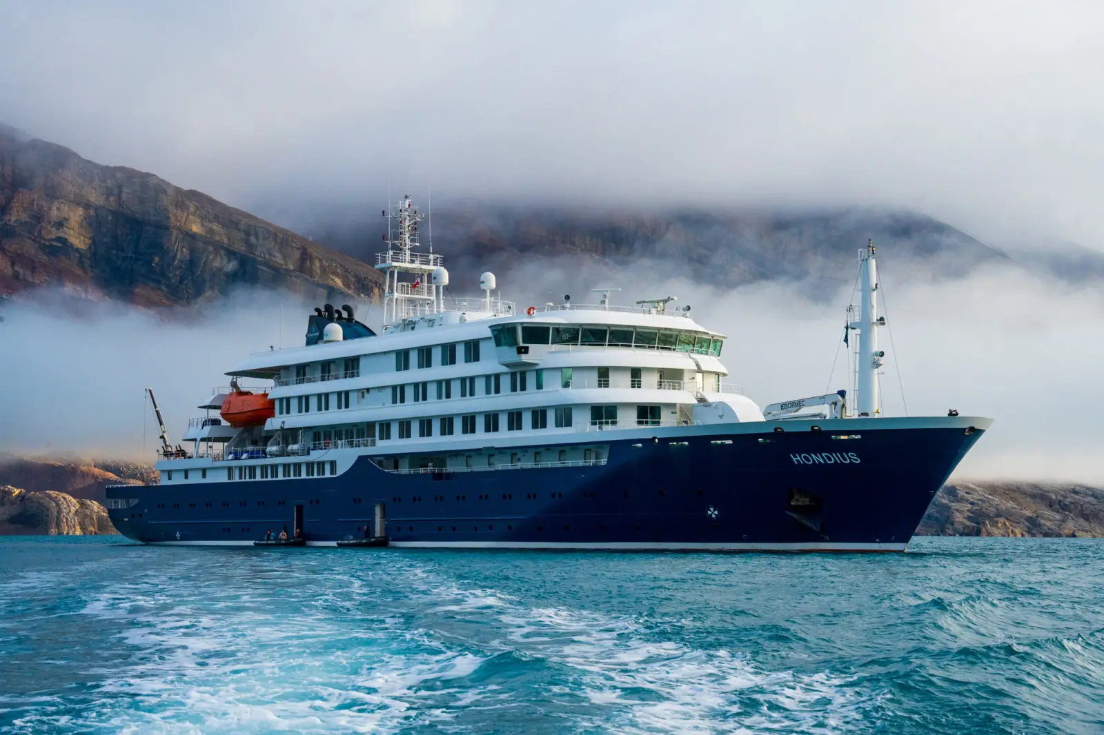 The MV Hondius, a dark blue and white expedition cruise ship operated by Oceanwide Expeditions, sailing on turquoise water in front of steep rocky cliffs partially covered by low cloud and mist