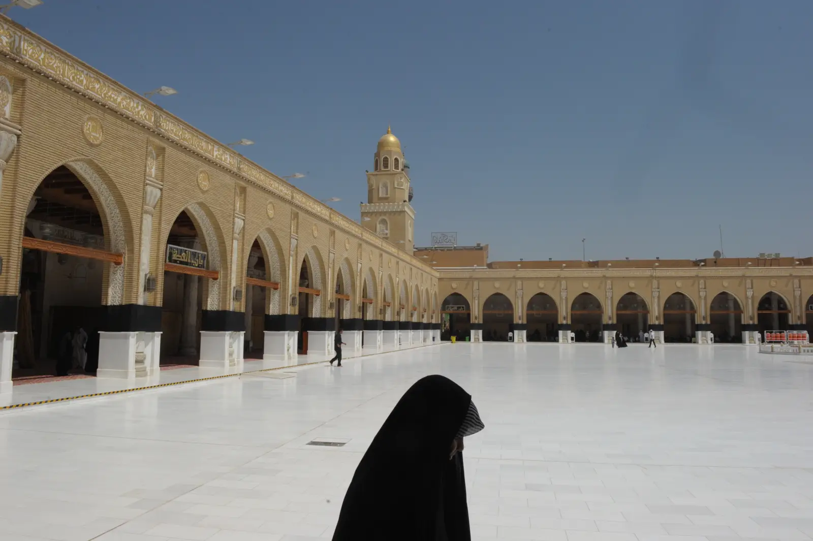 Wide courtyard of Kufa Mosque in Najaf, Iraq, with few visitors and a woman in black abaya walking under clear blue sky