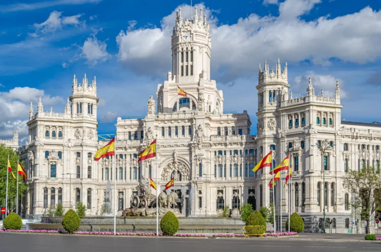 Palacio de Cibeles in Madrid with Spanish flags flying in front of the fountain and ornate white facade under a blue sky