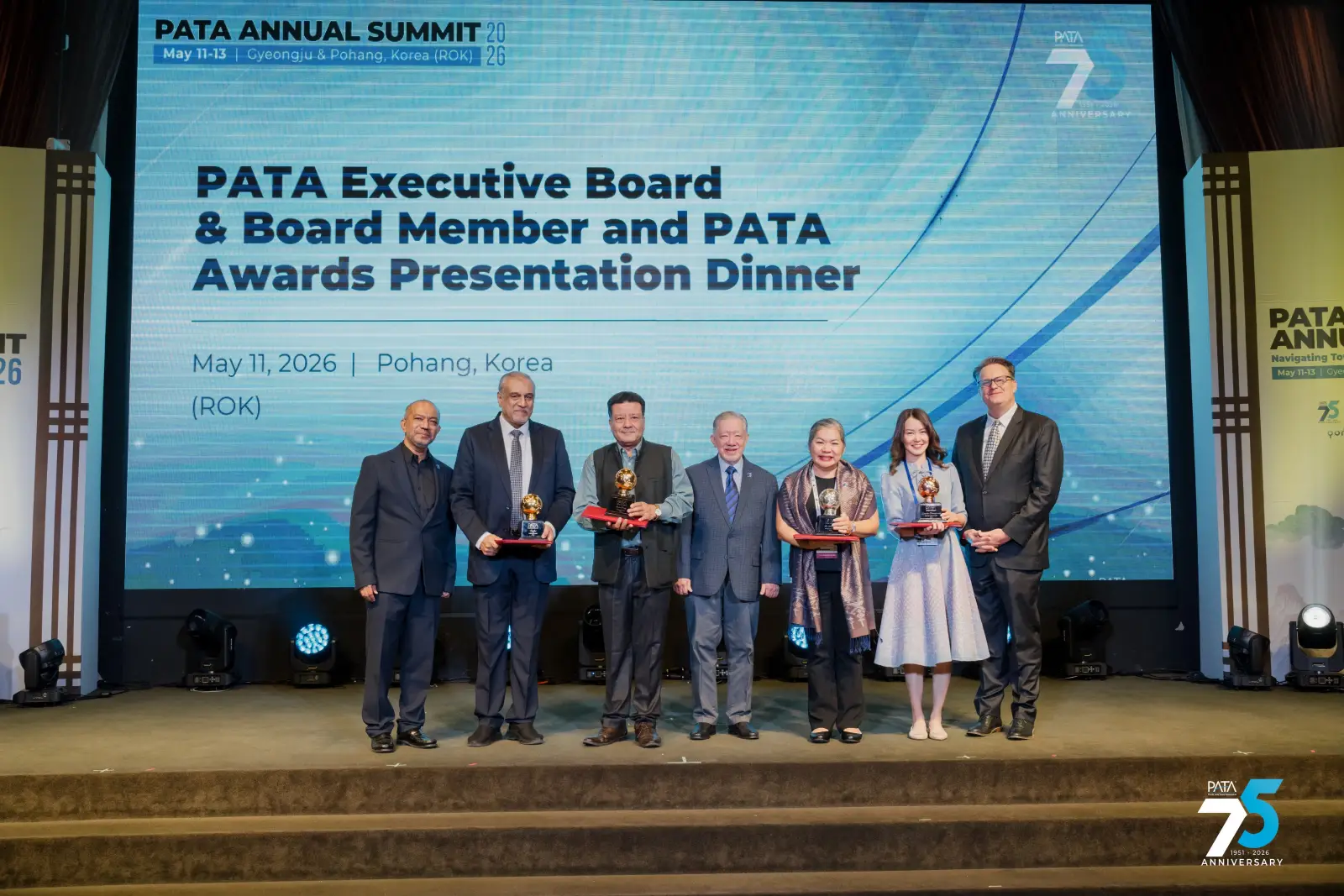 Six people stand on a stage at the PATA Executive Board and Board Member and PATA Awards Presentation Dinner on 11 May 2026 in Pohang, Korea. Four of them hold golden globe-shaped trophies and red presentation boxes. A large screen behind them displays the event name and date. The PATA 75th anniversary logo is visible in the lower right corner.
