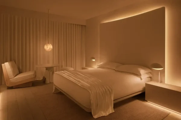A warmly lit hotel guest room at Public West Hollywood featuring a low-platform bed with white linen, a large illuminated headboard with amber backlight, a sculptural pendant light, a lounge chair, and fluted white wall panels, designed by Ian Schrager and John Pawson.