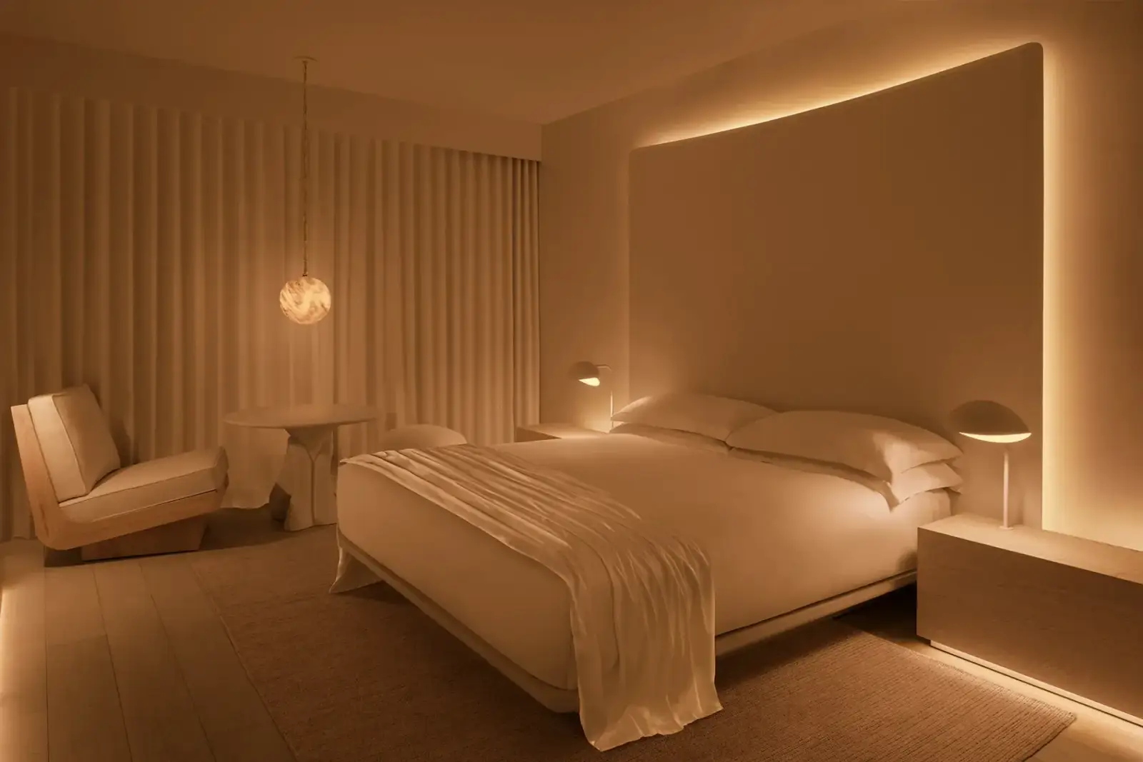 A warmly lit hotel guest room at Public West Hollywood featuring a low-platform bed with white linen, a large illuminated headboard with amber backlight, a sculptural pendant light, a lounge chair, and fluted white wall panels, designed by Ian Schrager and John Pawson.