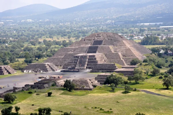 Aerial view of the Pyramid of the Moon at the ancient city of Teotihuacan, Mexico, seen from the top of the Pyramid of the Sun, with the Avenue of the Dead, smaller temple platforms, scattered visitors, and tree-covered valley and mountain range in the background.