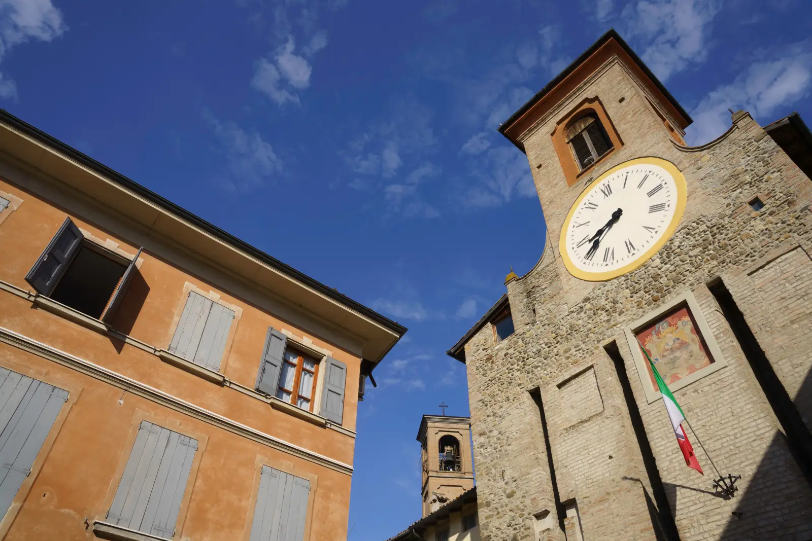 Low-angle view of a medieval stone clock tower with a large Roman numeral clock face and Italian tricolour flag displayed, alongside an ochre-coloured building with shuttered windows, in the historic centre of Reggio Emilia, Italy