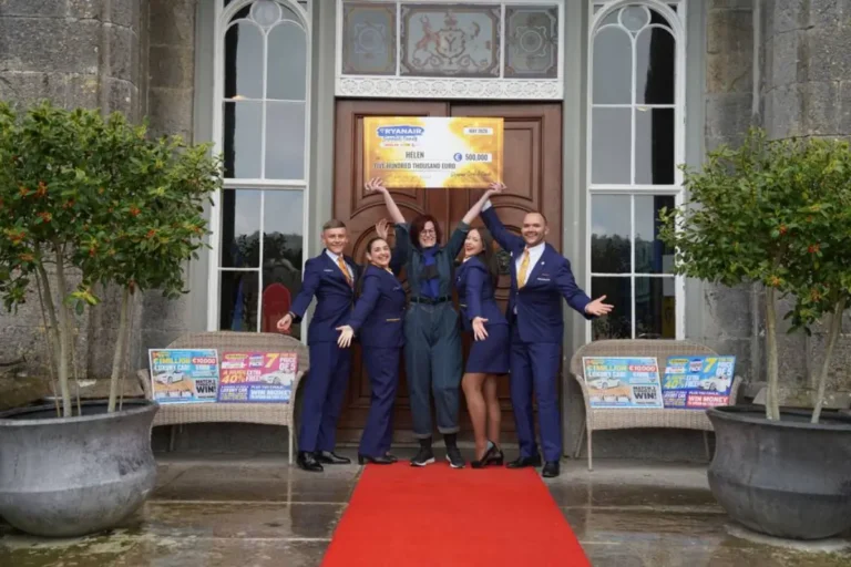 Aviation 28 Helen Swindells, dressed in dark casual clothing, stands at the entrance of Slane Castle in Ireland, raising a large ceremonial cheque for 500,000 euros above her head alongside four Ryanair cabin crew members in navy uniforms. A red carpet leads to the castle door, and Ryanair scratch card promotional displays are visible on either side.