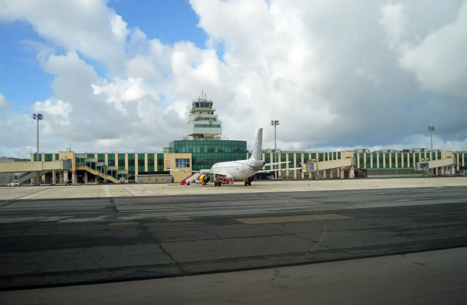 Ground-level view of Santiago de Compostela Rosalía de Castro Airport in Galicia, Spain, showing a Vueling aircraft parked on the apron in front of the terminal building with the air traffic control tower and Santiago signage visible against a partly cloudy sky