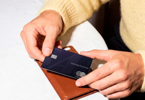 Close-up of two hands holding a dark navy metal SAS Business Executive Mastercard card above a brown leather passport wallet on a white surface