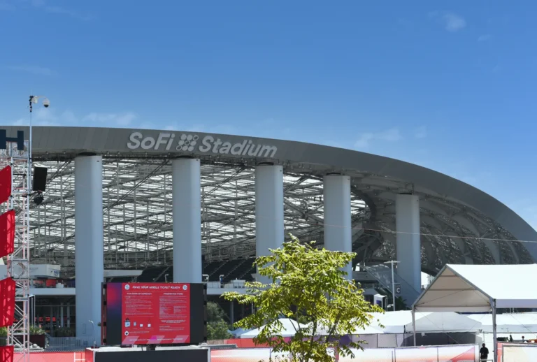 Exterior daytime view of SoFi Stadium in Inglewood, California, showing the venue's distinctive curved roof structure supported by large white columns under a clear blue sky.