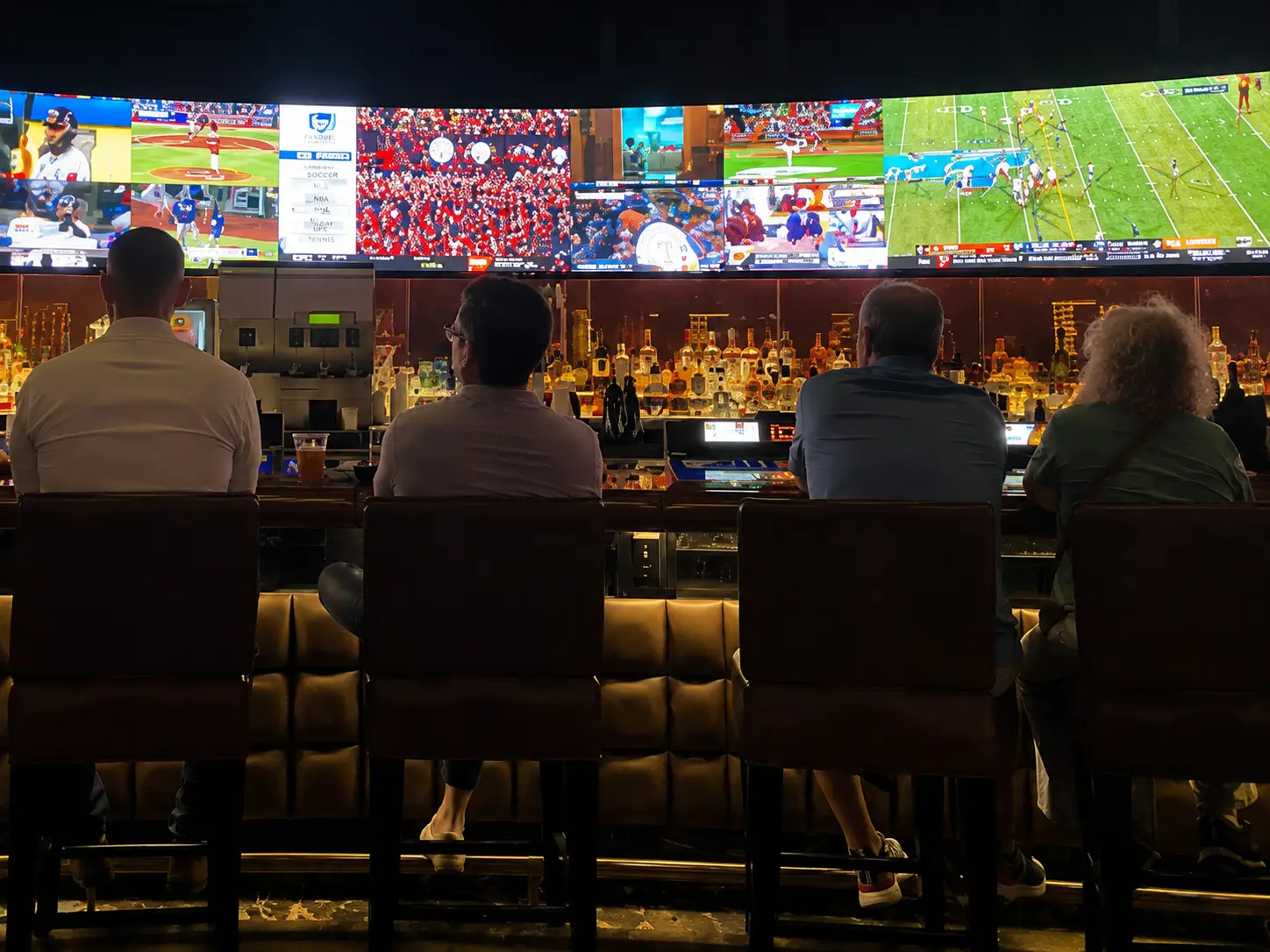 People sitting at a sports bar watching multiple live baseball and football games on large screens above the bar counter at night.