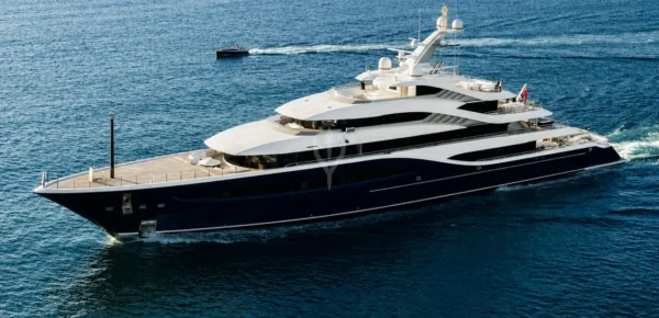 Aerial view of the 88-metre superyacht Angelique by Turquoise Yachts cruising at sea, showing its dark navy steel hull, white aluminium superstructure and multiple deck levels with a tender boat visible in the background