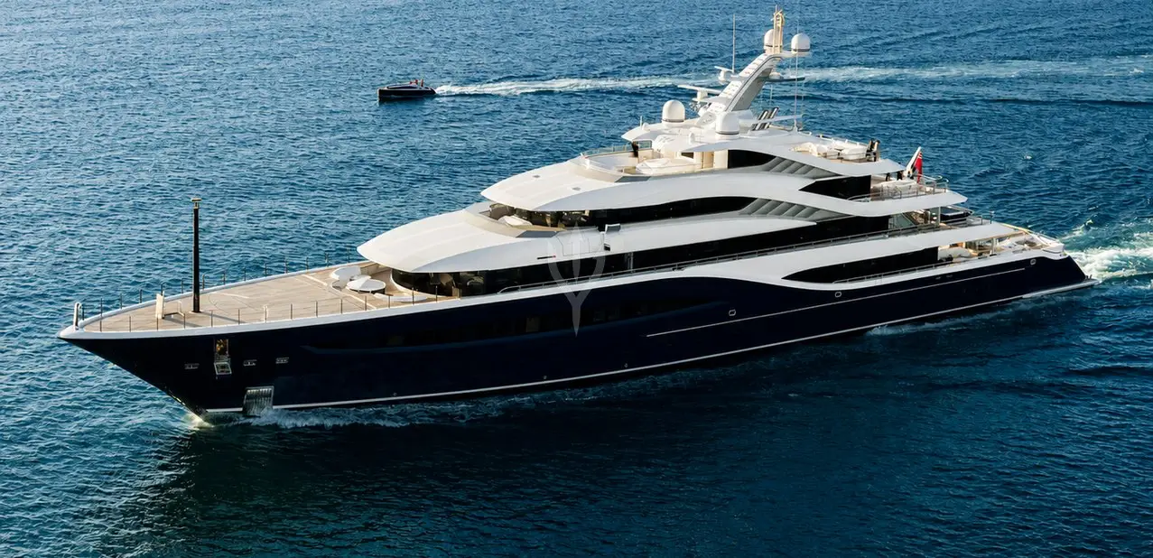 Aerial view of the 88-metre superyacht Angelique by Turquoise Yachts cruising at sea, showing its dark navy steel hull, white aluminium superstructure and multiple deck levels with a tender boat visible in the background