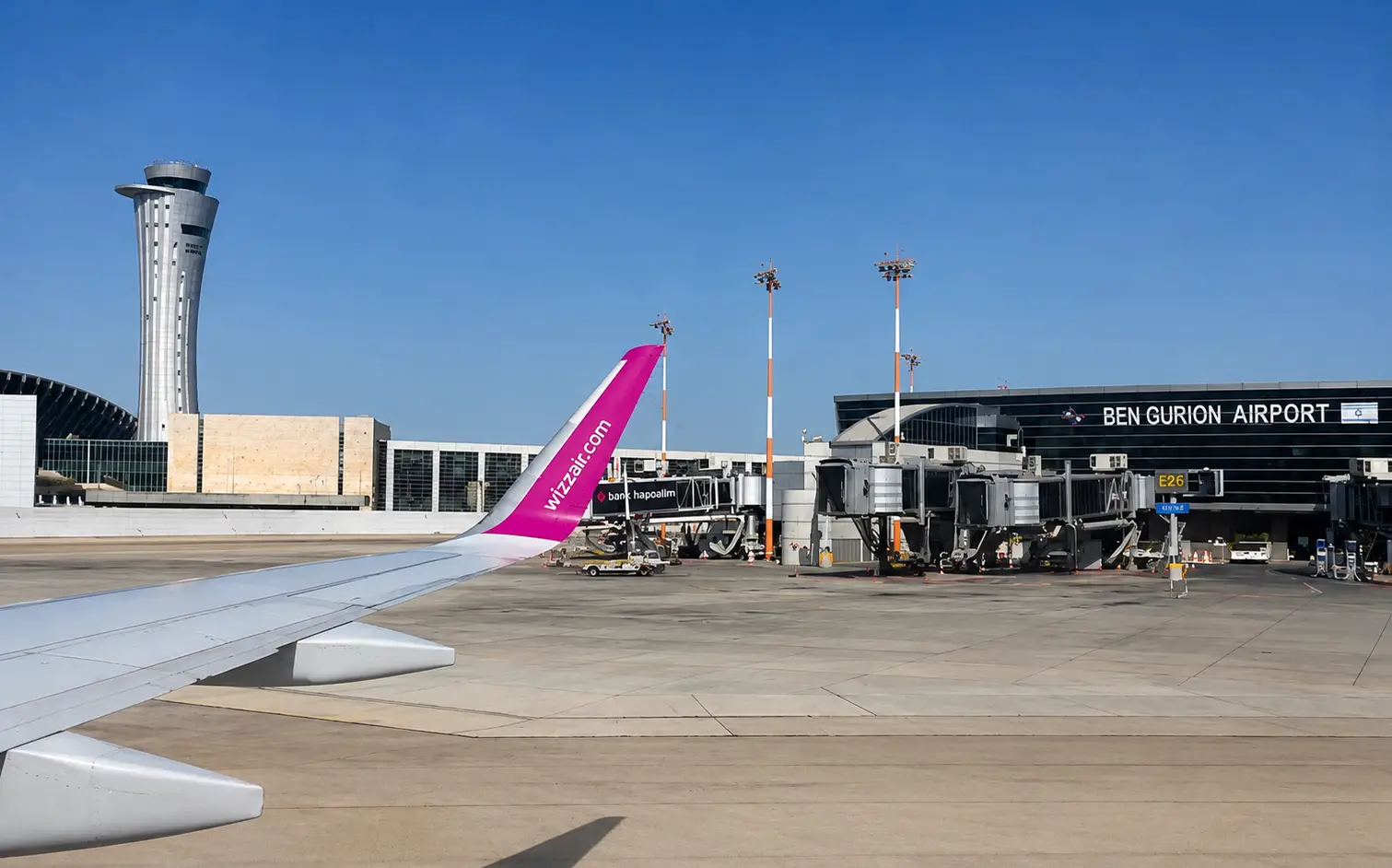 Wizz Air aircraft wing at Ben Gurion Airport in Tel Aviv, Israel, beside the terminal building under a clear blue sky.