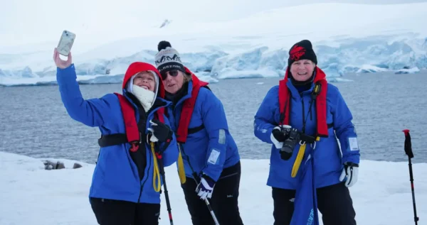 Women travelers in blue expedition jackets take a selfie on the snowy Antarctic coastline during an Aurora Expeditions cruise.