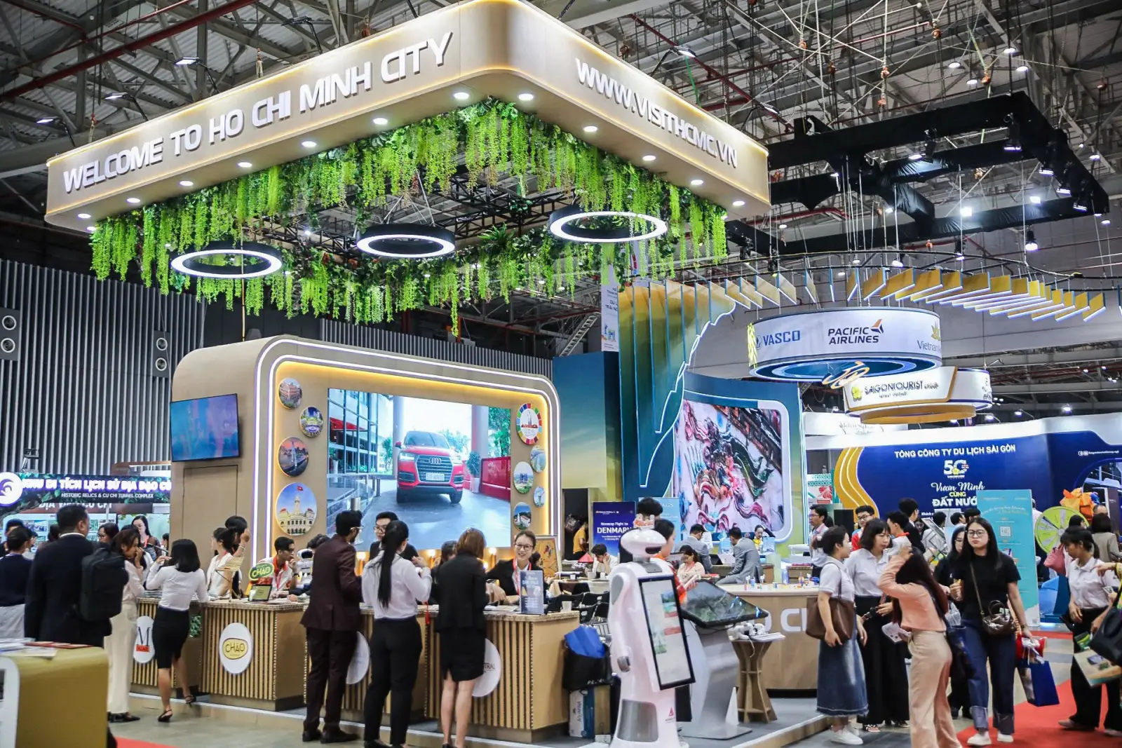 Ho Chi Minh City tourism pavilion at a travel trade fair, featuring a decorative green hanging plant display, digital screens, and branded booths from Pacific Airlines, VASCO, and Saigontourist, with visitors browsing the exhibition floor