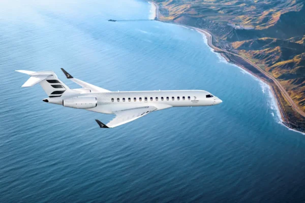 Aerial view of a Bombardier Global 8000 business jet in white livery cruising over a blue sea and coastal mountain terrain