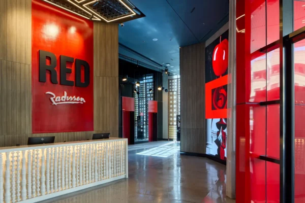 Home 8 Lobby and reception area of Radisson RED Cebu Mandaue, featuring a bold red wall with the RED Radisson logo, wood panelled accents, a white textured front desk, geometric ceiling lighting and a corridor leading to red toned digital art installations and elevators.